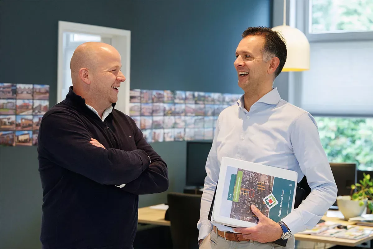 Twee medewerkers van Ditters Makelaars Arnhem in gesprek op kantoor, met dossiers en woningpresentaties op de achtergrond.